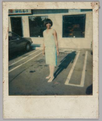 [Woman in light blue top and skirt and white stockings and shoes standing in the parking lot of a building with a "Deli" sign to the left]