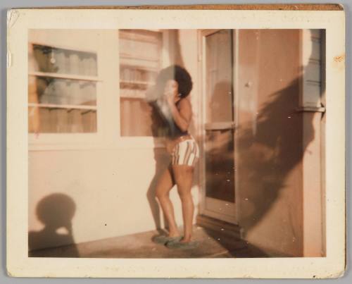 [Woman outside a house and smoking, she is leaning by the window and wearing striped shorts and a black tank top]