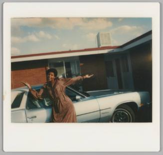 [Woman in dress posing with her arms spread wide in front of a parked car, one storey building behind her]