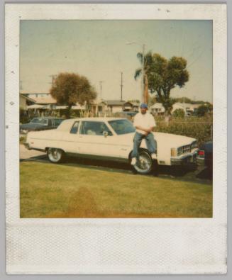 [Man sitting on the hood of a white car parked in a driveway by front lawn, there are houses and trees in the distance]