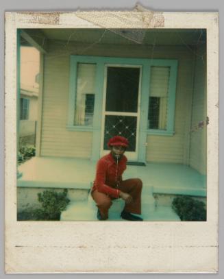 [Man in red shirt, pants and hat sitting on the steps of a house with a blue deck and blue trim around the door and windows]