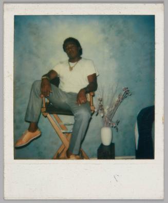 [Man in yellow shoes sitting in director's chair with his legs spread wide, there is a vase below him to the right]