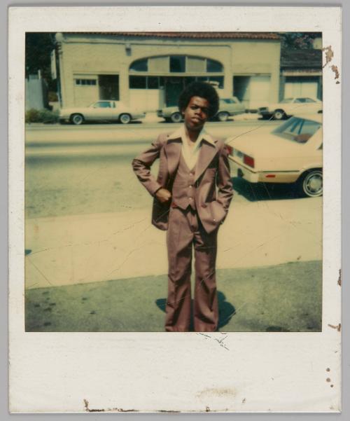 [Young man in a large brown three-piece suit and a white shirt with a wide collar, he is standing on the grass by a sidewalk, cars are parked behind him]