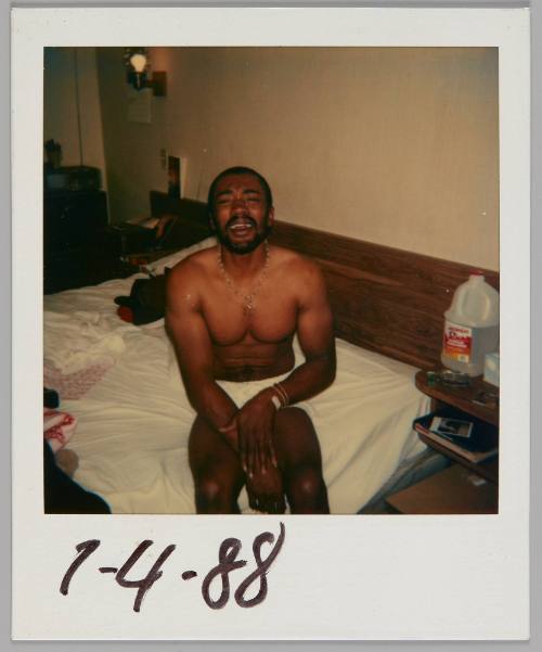7-4-88 [Man in underwear and necklace sitting on the side of the bed and looking up at the camera, plastic jug of water on bedside table]