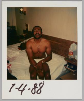 7-4-88 [Man in underwear and necklace sitting on the side of the bed and looking up at the camera, plastic jug of water on bedside table]