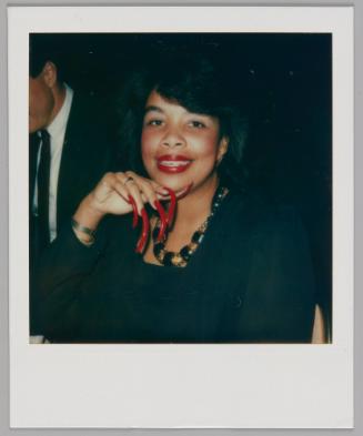 [Woman with long red nails and a black and gold necklace resting her chin on her hand and smiling]