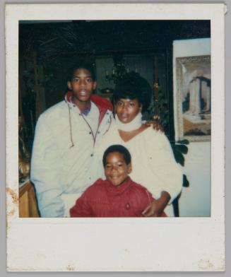 [Man in light-coloured jacket with red lining standing with his arm around a woman in a neck brace, boy in red jacket in front of them and smiling]