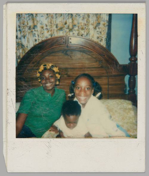 [Young woman with yellow curlers in her hair and wearing a green polo shirt sitting on a bed with an older girl and baby, the baby is looking down]