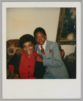 [Boy in grey three-piece suit with red tie a pocket square leaning on the arm of a chair with his arms around a woman in a red shirt with black buttons]