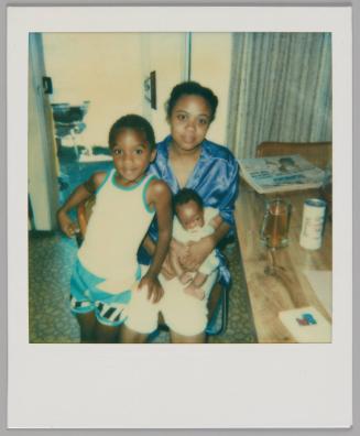 [Woman in shiny top sitting with baby on her lap, boy in white tank top and blue shorts standing beside her with his hand on her knee]