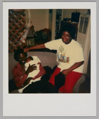 [Man sitting on couch holding a sleeping baby against his shoulder, woman in red pants sitting on arm of couch]