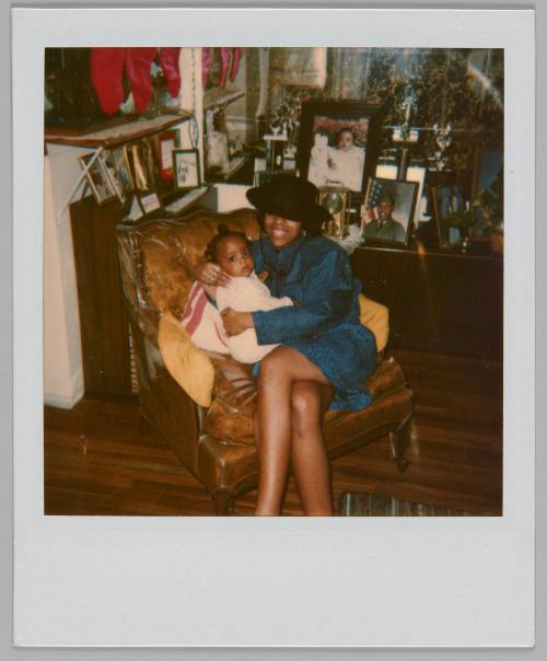 [Woman in blue dress and black hat sitting on brown armchair with a baby, there are red stockings hanging on left and framed family photographs on the tables]