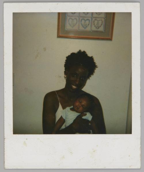 [Woman with short hair and white tank top sitting down and holding sleeping baby wearing a t-shirt, there is a picture frame on the wall behind them]