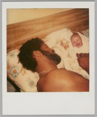 [Shirtless man lying on bed  with floral pillow cases and tickling a baby wrapped up in a blanket, the baby is laughing]