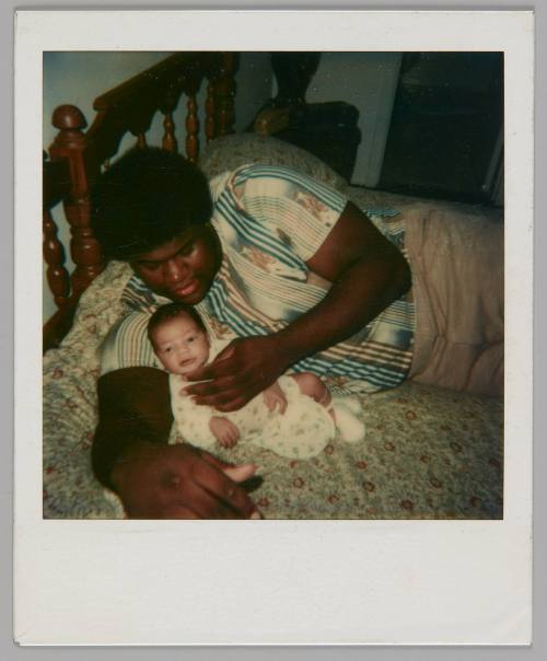 [Man in striped short-sleeved shirt holding and looking down at a baby, they are lying on a bed with a wood headboard]