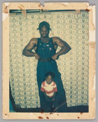 [Man in denim overalls and no shirt in bodybuilder pose with boy standing between his legs, behind them is a backdrop of American money bills]
