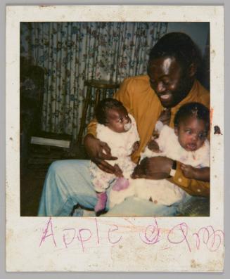 Apple [illeg.] [Man in yellow shirt holding two babies and smiling down at them, baby on left has pacifier in mouth]