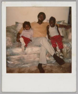 [Man in yellow t-shirt with a cigarette in his mouth and his arms around two girls, one on either side of him, they are all sitting on couch wrapped in plastic]