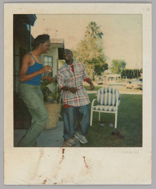 [Two people standing outside house with a blue and white striped patio chair on the lawn, man on right in white, red, and blue plaid shirt]
