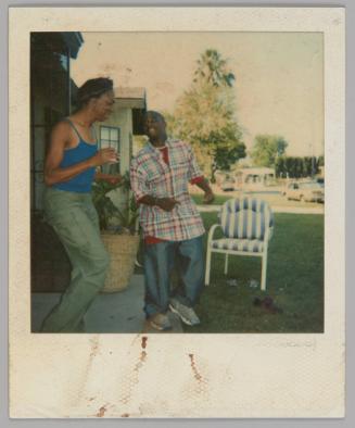 [Two people standing outside house with a blue and white striped patio chair on the lawn, man on right in white, red, and blue plaid shirt]