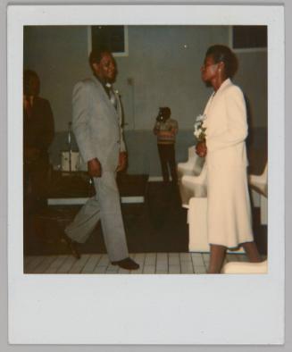 [Man in grey suit walking towards woman in white dress holding a bouquet, they are inside a room with high windows and white benches]