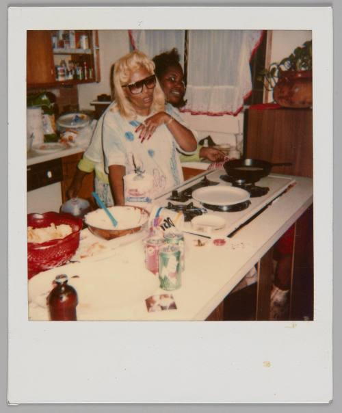 [Blonde woman in sunglasses standing behind kitchen counter with baking supplies out, behind her there is someone peeking out and smiling]