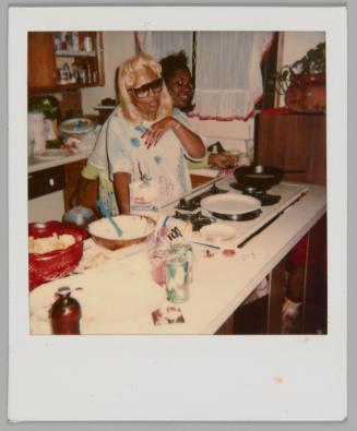 [Blonde woman in sunglasses standing behind kitchen counter with baking supplies out, behind her there is someone peeking out and smiling]