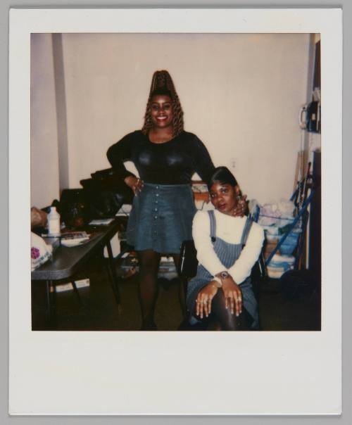 [Woman in black shirt and denim skirt standing with one hand on her hip and other hand on the woman sitting down beisde her, woman sitting has her hands crossed over her knees]