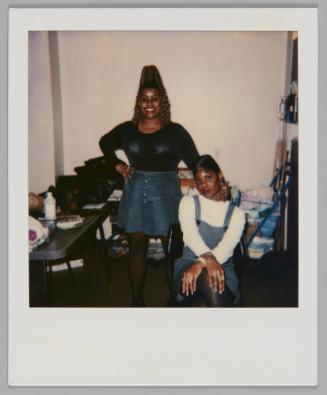 [Woman in black shirt and denim skirt standing with one hand on her hip and other hand on the woman sitting down beisde her, woman sitting has her hands crossed over her knees]