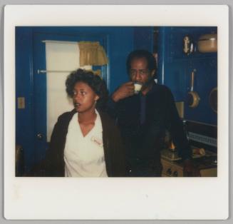 [Woman in white shirt and cardigan with name tag standing beside man sipping from a small cup, they are in a blue kitchen]