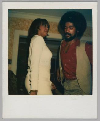 [Woman in white sweater and pants standing at looking at man in red shirt and open vest, he is looking downward]