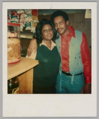 [Woman in blue knit vest standing by man in red shirt with an open denim vest, he has his arm around her, there are bags of snacks behind them]