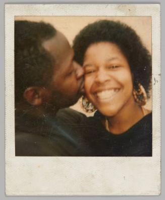 [Close-up of man kissing woman on the cheek, she is wearing gold hoop earrings and a black shirt]