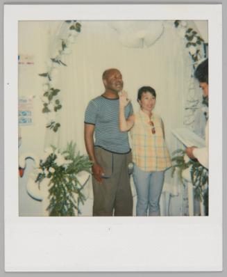 [Man and woman standing under a wedding arch covered in white flowers with a swan statue to the left, woman in yellow plaid sleeveless top and is holding her hand up]