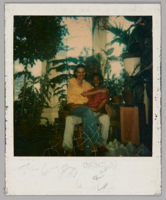 7 - 6 - 80 [illeg.] [Man with moustache and yellow shirt sitting on lap of man in red shirt, they are in a room with large windows and many potted plants]