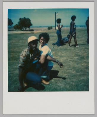 [Man in white hat with black brim and woman in white t-shirt kneeling on grass with their arms linked,  other people are walking by]
