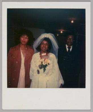 [Woman in wedding dress and veil with bouquet of daisies standing between woman in red coat and man in suit]