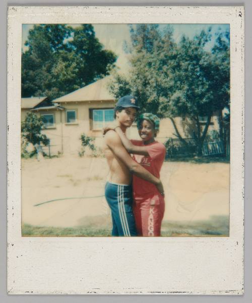 [Shirtless man in baseball cap and track pants hugging woman in a pink t-shirt and pants with blue curlers in her hair, they are standing outside a house]