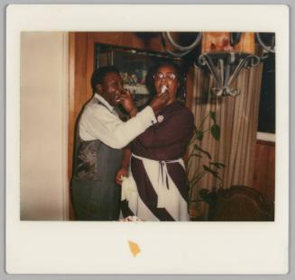 [Man and woman stuffing each others mouth with cake, man is wearing a waistcoat and the woman is wearing glasses and a striped skirt]