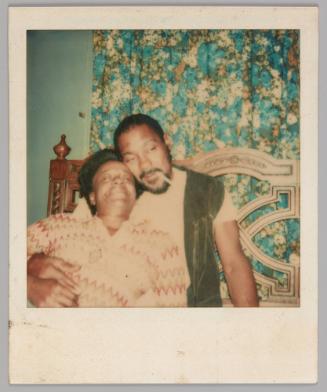 [Man in white t-shirt and black vest with a cigarette in his mouth holding the person reclining on him, there is a blue patterned curtain behind them]