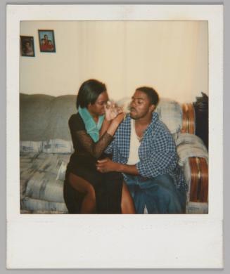 [Woman and man sitting on grey couch with their arms intertwined and each taking a sip from a wine glass, there are family portraits on the wall behind them]