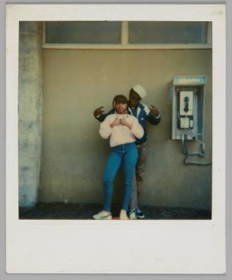 [Teenage boy and girl posing beside a phonebooth, girl is standing in front in blue jeans and a pink sweatshirt and the boy is standing behind her in a white hat]