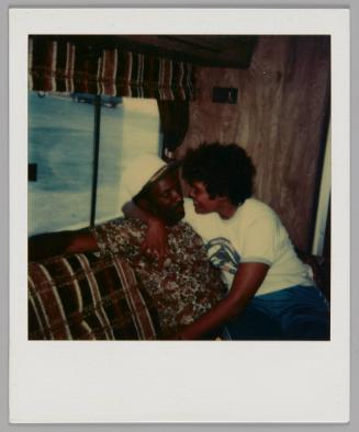 [Man in white hat with black brim and patterned short-sleeved shirt sitting on striped couch with a woman sitting on his lap and hugging him]