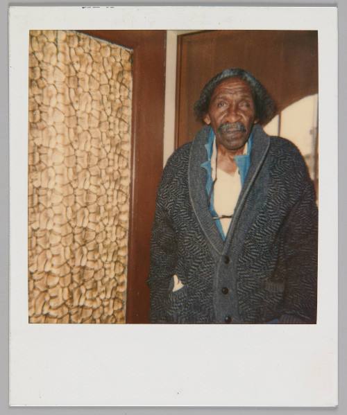 [Older man with moustache wearing a knit cardigan and his glasses around his neck, he is standing beside a patterned curtain]