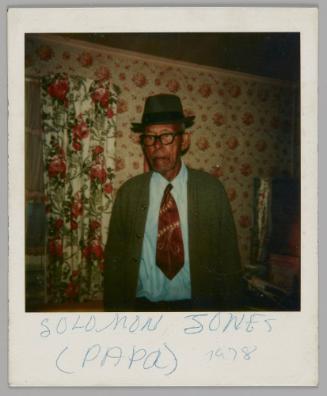 Solomon Jones (Papa) 1978 [Older man in fedora hat, black rimmed glasses, a blue shirt and red tiestading in a room with floral wallpaper and curtains]