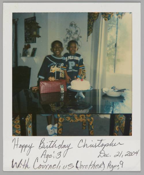 Happy Birthday Christopher, Age:3, with Cornelius (brother) Age: 9 Dec. 21, 2004 [Two boys wearing blue sports jerseys standing behind glass dining table with white round birthday cake]