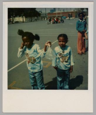 [Two girls standing on paved playgound wearing matching tiger tops, girl on left is holding plastic bag filled with water and a fish]