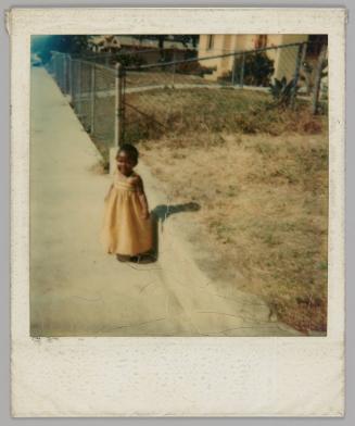 [Young girl in dress standing on the sidewalk by a gress lawn, chainlink fence behind her]