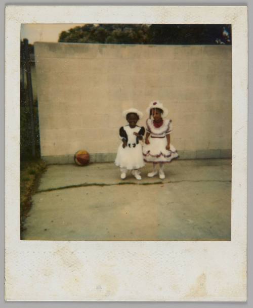 [Two small girls in frilly white dresses and white hats, standing outside on pavement and in front of a cement wall]