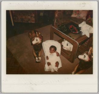 [Baby in white outfit sitting in a white baby carrier on a brown carpet, baby is surrounded by large ceramic dogs]